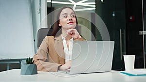 Pensive woman sitting at desk in office while typing on portable laptop