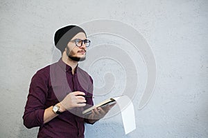 Pensive writer man holding a notebook and pencil. Bearded and dressed with shirt and black hat, wearing glasses.