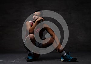 Pensive young African-American basketball player in sportswear sitting on a ball over dark background.