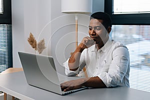 Pensive young African-American businessman working on business strategy thinking looking to laptop screen sitting at