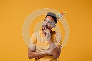 Pensive young african american guy in casual t-shirt, birthday hat posing isolated on yellow orange wall background