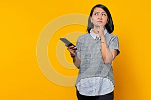 Pensive young Asian woman using mobile phone with fingers on chin ang lookin aside over yellow background