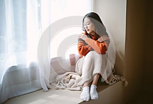 Pensive young Asian woman in warm knitted clothes sitting on windowsill in room. Negative human facial expression