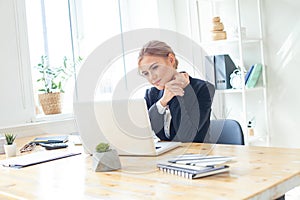 Pensive young beautiful businesswoman working on laptop and keeping hand on chin while sitting at her workplace