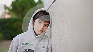 Pensive young boy in gray hoodie leaning against a wall outdoors