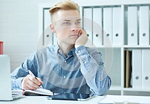 Pensive young business man sitting at the dask with laptop and looking at the window.