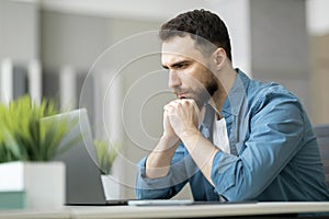 Pensive Young Businessman Sitting At Workplace And Looking At Laptop Screen