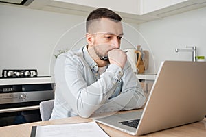 Pensive young Caucasian man sit at table at home kitchen work online on laptop pondering thinking. Thoughtful millennial