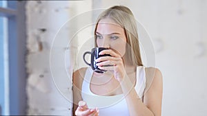 Pensive young European woman looking on window drinking hot tea at white loft background