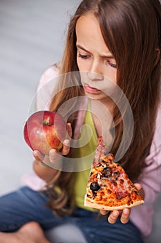 Pensive young girl ponder on choosing a fruit or a slice of pizza as a snack