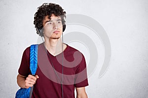Pensive young guy with headphones on head and blue rucksack, wearing maroon casual t shirt, listens to music, looking thoufhtfully