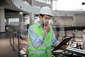 Pensive young islamic engineer builder touches his beard in protective clothing and hard hat looks at tablet
