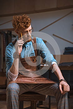pensive young male fashion designer in apron and eyeglasses sitting and thinking with pencil