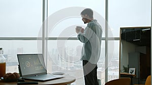 Pensive young man drinking coffee looking at city from apartment window