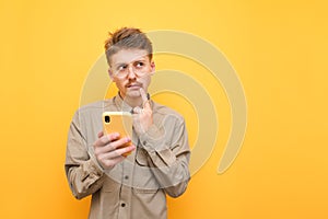 Pensive young man in glasses and shirt stands with smartphone in hand against yellow background, looks away and thinks. Thoughtful