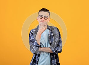 Pensive young man in glasses touching his chin, thinking over something on orange studio background