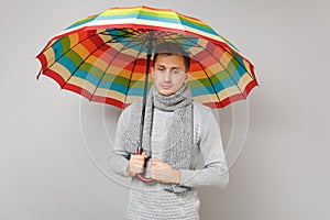 Pensive young man in gray sweater, scarf looking down, holding colorful umbrella on grey background. Healthy