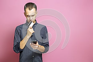 Pensive young man holding card and smartphone on pink background.