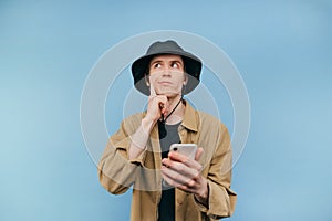 Pensive young man in panama and shirt with a smartphone in his hand stands on a blue background, looks away and thinks