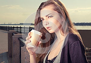 Pensive young trendy woman drinking take away coffee and standing leaning back granite fence urban scene.