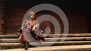 Pensive young ukrainian cossack dressed traditional clothing holding saber in his hand sitting on the steps