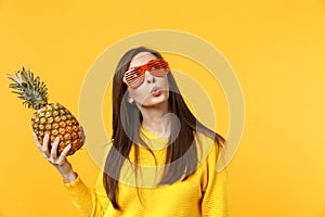 Pensive young woman in funny glasses looking up, holding fresh ripe pineapple fruit isolated on yellow orange background