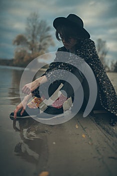 Pensive young woman in hat sits at beach next to lake and touche