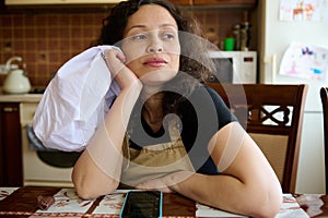 Pensive young woman housewife, baker confectioner in chef's apron, sitting at table in home kitchen, looking away