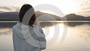 Pensive young woman with long hair seen from behind looks at the lake at sunset