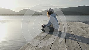 Pensive young woman with long hair seen from the side looks at the lake at sunset sitting on an old wooden pier