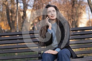 Pensive young woman with long wavy brown hair sits in park on bench and talks on phone and gesticulating