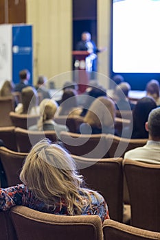 People Attending Business Conference Sitting in Front of the Host Speaking on Stage Against Big Screen.