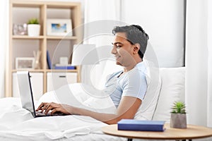 Happy indian man with laptop in bed at home