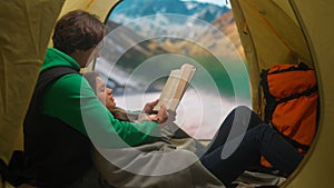 People on campsite traveling and hiking, exploring nature. Young loving couple reading a book inside the tent at lake