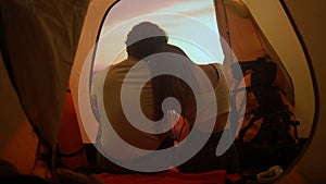 People on campsite traveling and hiking, exploring nature. Young loving couple sitting inside the tent looking at sunset