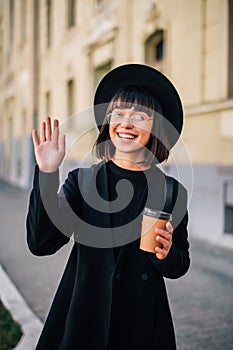 People and drinks concept. Happy young woman drinking takeaway coffee from paper cup over and waving hand on city street