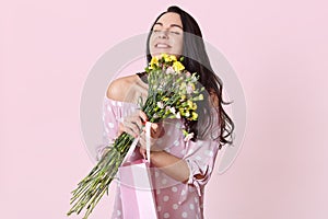 People, enjoyment and happiness concept. Positive dark haired woman with black hair, embraces flowers, carries gift bag, poses