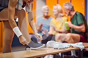 People in gym locker room. Young woman tying her shoe, seniors in the backgorund sitting and talking