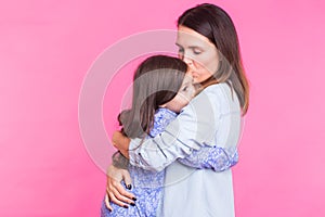 People, happiness, love, family and motherhood concept - happy little daughter hugging and kissing her mother over pink