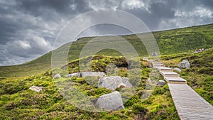 People hiking on wooden boardwalk, between boulders and heathers, leading to Cuilcagh Mountain