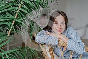 People, leisure concept. Beautiful European woman dressed in stylish clothes, sits on wooden chair near green plant, looks positiv