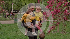 People in park. Dad hold 2 baby daughter girl in arms near blossoming apple tree and sniffing flowers. Parents and fun