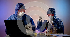 People in protective clothing with hoods, respirators, glasses and gloves sit at a table with a laptop and test tubes