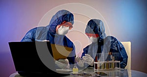 People in protective clothing with hoods, respirators, glasses and gloves are sitting at a table with a laptop. one