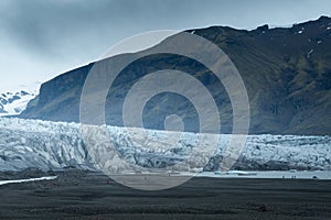 People in red and blue outdoor jackets dwarfed by a glacier on a cold, cloudy day in southern Iceland.