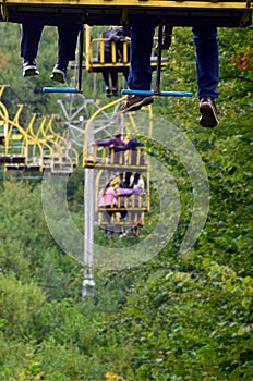 People ride on a cable car. The legs of passengers hang over the mountain forest