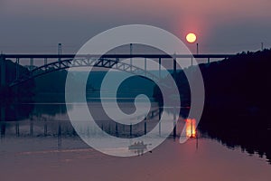 People are sailing on a boat on the river, there is a railway bridge and the sun over the river