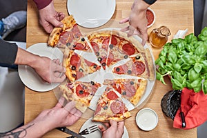 People shares fresh pizza with pepperoni sausage and black olives on wooden table in restaurant.