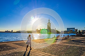 People are sitting on bench at promenade embankment of Lake Malaren looking at Stockholm City Hall Stadshuset