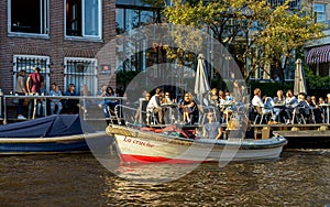 People sitting in Cafe restaurant on the canal in Amsterdam with parked small city tour boat, the Netherlands, October 13, 2017